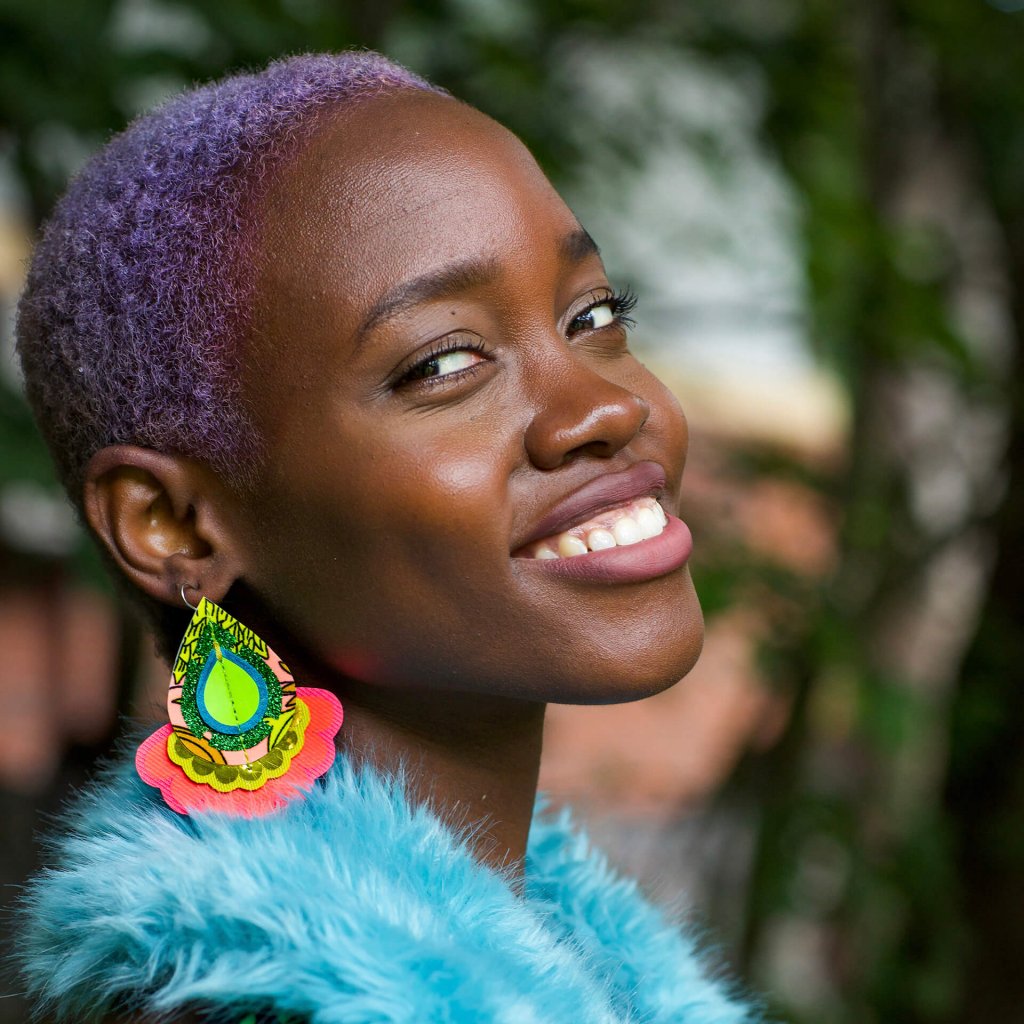 A young black woman with cropped lilac hair wears a pair of colourful, oversize statement earrings and a bright blue faux fur wrap. She is smiling and looking sideways, over her shoulder, into the camera.