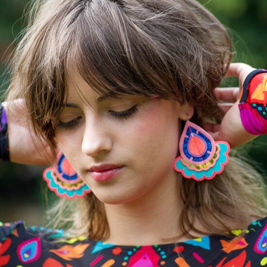 A young white woman with shaggy brown hair is looking down and holding her hair back to reveal her colourful statement earrings. She is wearing a colourful, patterned, long sleeve top which is just visible