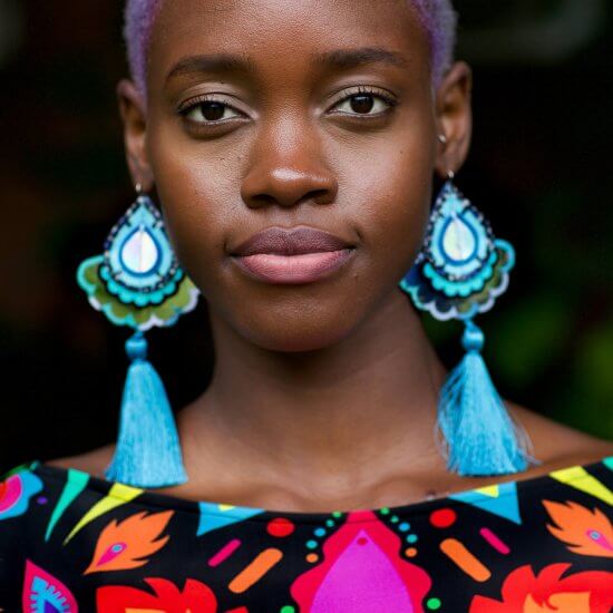A black female model stares into the camera. She wears a pair of large silky tassel earrings with bright blue tassels and a colourful, patterned lycra bodysuit