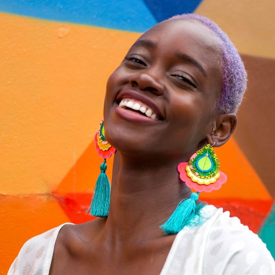 A black woman with short lilac hair wearing large colourful tassel earrings is tilting her head backwards and smiling. Behind her is a colourfully painted yellow, orange and blue wall.