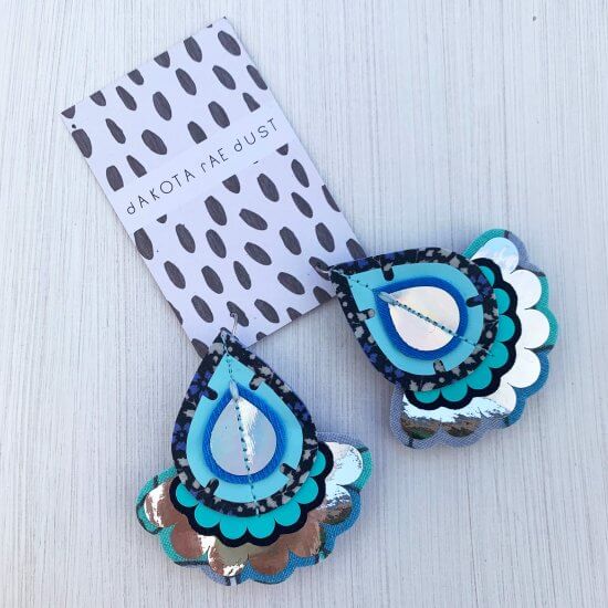 A pair of teardrop shaped, statement oversize earrings in multiple shades of blue and silver are seen mounted on a black and white patterned dakota rae dust branded card, against an off white background