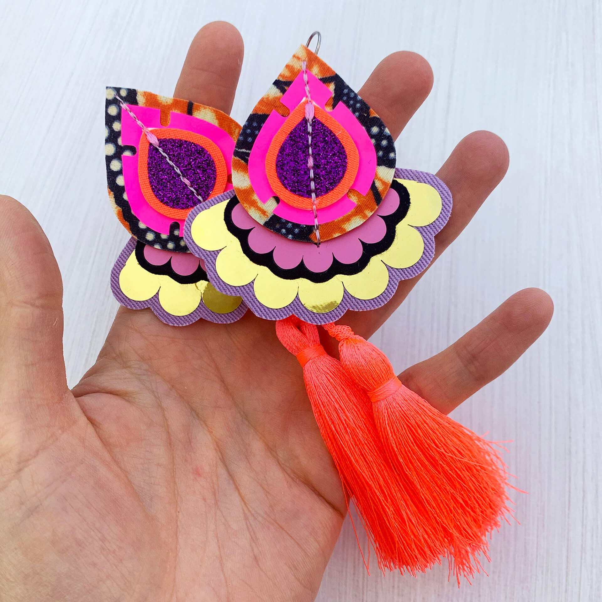 A pair of colourful decorative teardrop shaped earrings with neon coral silky tassels are held in a woman's open hand against an off white background