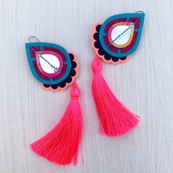 A pair of decorative teardrop shaped, neon pink tassel earrings mounted on a black and white patterned dakota rae dust branded card, photographed against an off white background