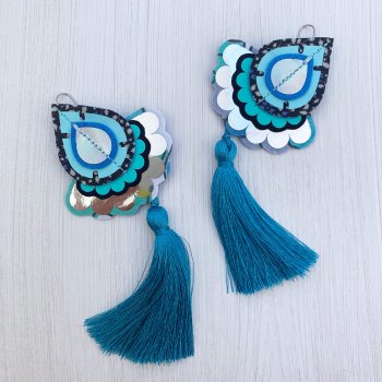 A pair of decorative teardrop shaped statement tassel earrings in shades of blue and silver mounted on a black and white patterned dakota rae dust branded card, photographed against an off white background