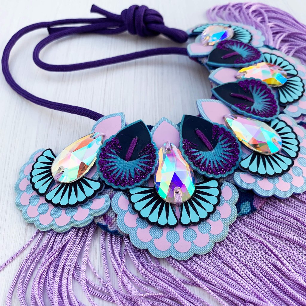 A lilac, light blue and purple statement jewel bib necklace with a lilac fringe photographed against an off white background
