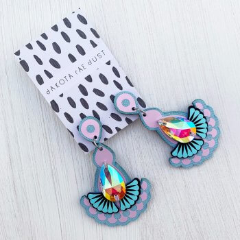 A pair of pale blue and lilac, fan shaped Statement jewel earrings adorned with an iridescent gem are seen mounted on a black and white patterned, dakota rae dust branded card against an off white background