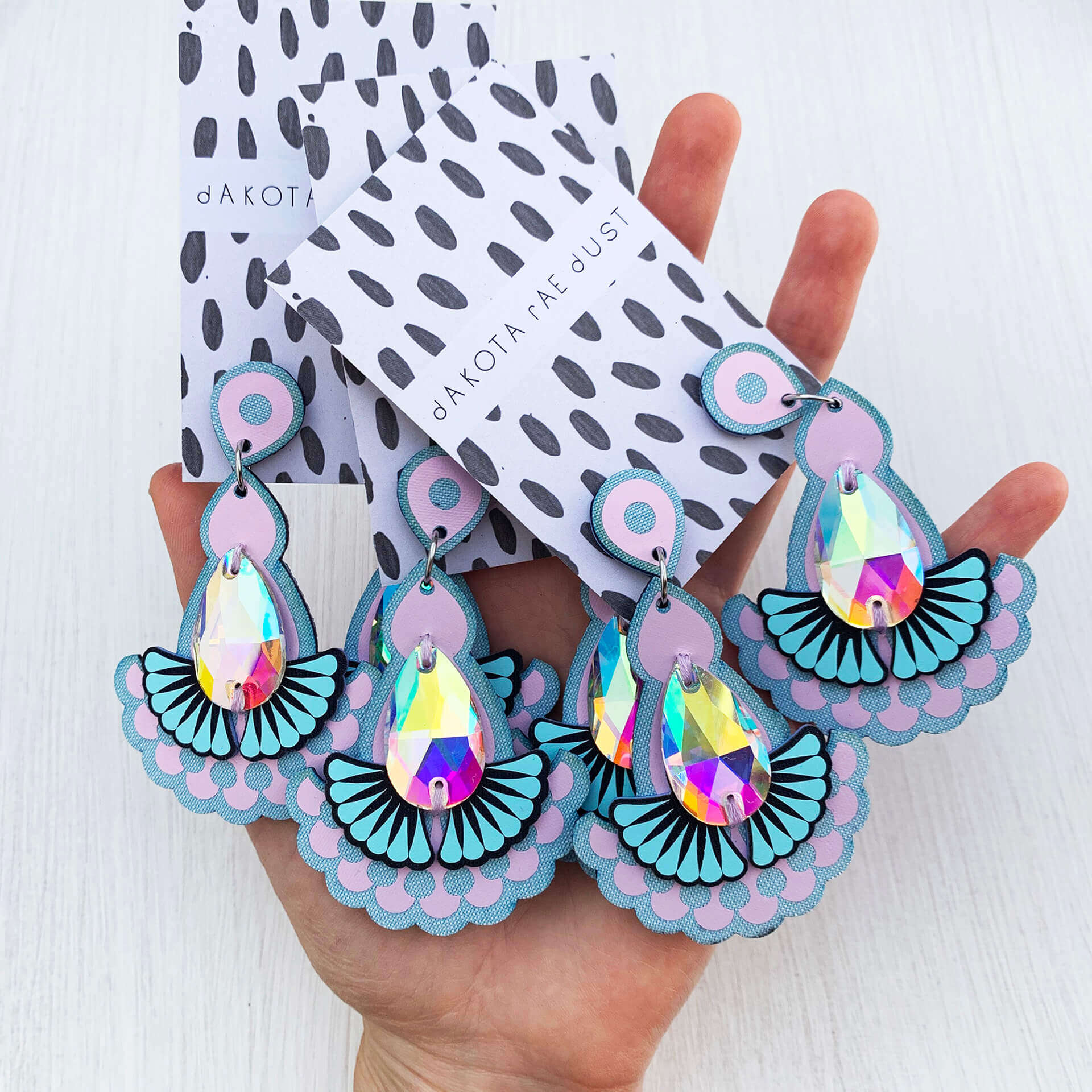 Several pairs of the same laser cut lilac and light blue jewelled statement earrings mounted on dakota rae dust branded cards are held in a woman's open hand