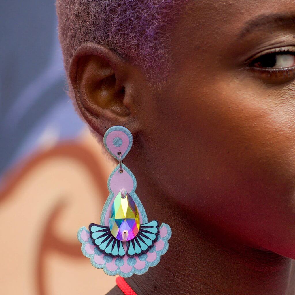 close up of a young black woman with short lilac hair wearing an ornate teardrop shaped statement jewel earrings. The side of her face and ear are in focus, the grey blue and peach block colour background os blurred.