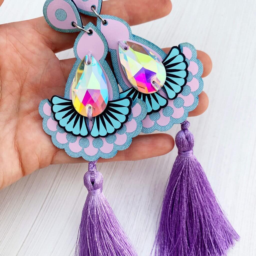 A pair of lavender, lilac and light blue statement jewel tassel earrings, featuring silky tassels and iridescent gems held in a white woman's hand against an off white background