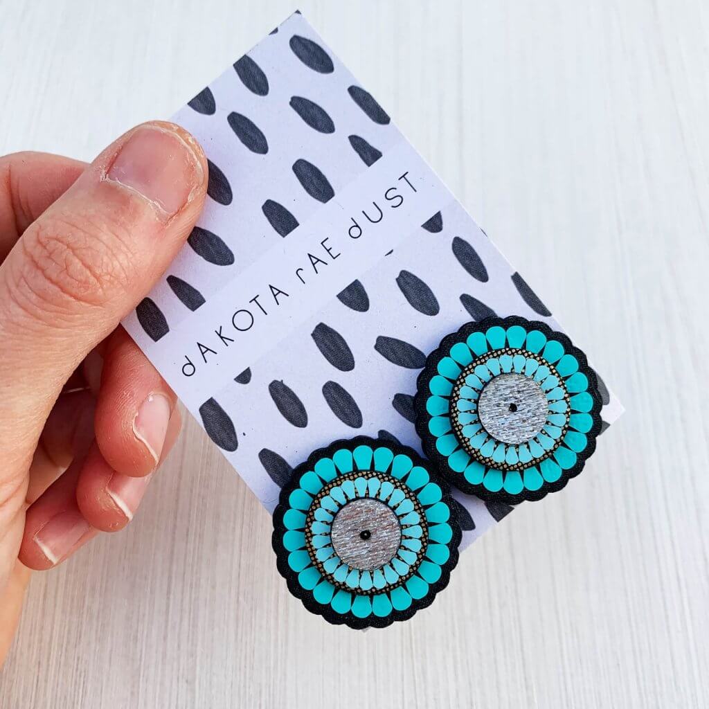 A pair of laser cut ornate and oversize turquoise stud earrings mounted on a black and white patterned dakota rae dust branded card held in a white woman's hand against an off white background
