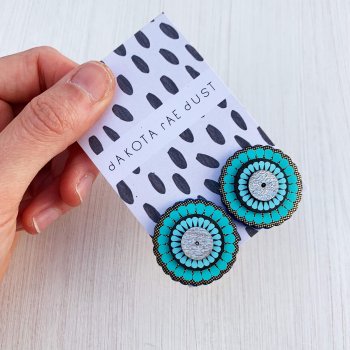 A pair of oversize turquoise gold studs mounted on a black and white patterned, dakota rae dust branded card held in a white woman's hand against an off white background