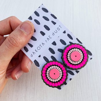A pair of oranate and oversize studs mounted on a black and white patterned, dakota rae dust branded card are held in a white woman's hand against an off white background