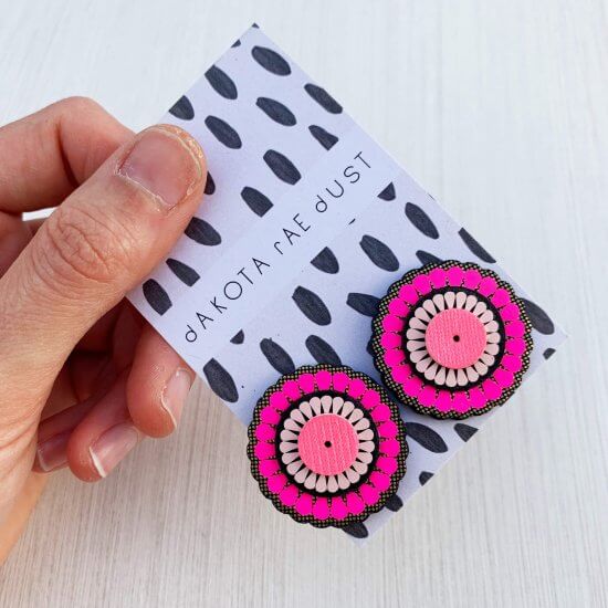 A pair of oranate and oversize studs mounted on a black and white patterned, dakota rae dust branded card are held in a white woman's hand against an off white background