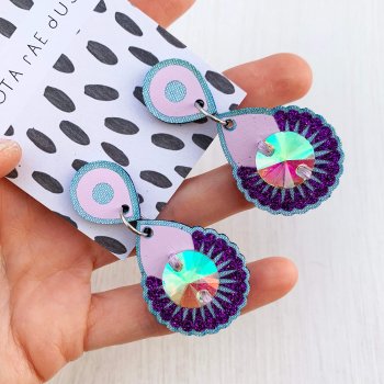 A pair of lilac and light blue dangly jewel earrings mounted on a black and white patterned, branded dakota rae dust card and held in a white woman's hand