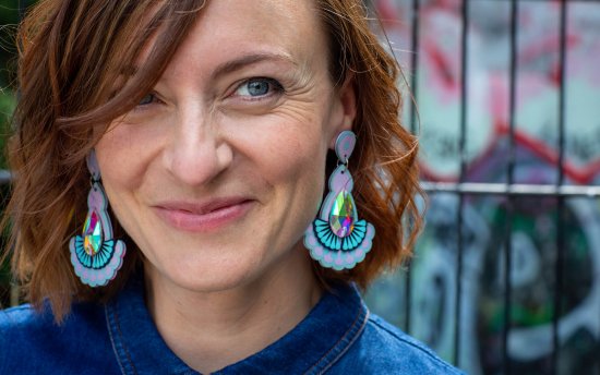 A woman with short brown hair is looking beyond the camera and smiling. She is wearing a pair of lilac and light blue statement jewel earrings and a denim shirt. Behind her is a background of out of focus street art in blues, turquoise and red.