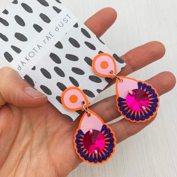 A pair of bright orange, pink and lilac dangly jewel earrings, mounted on a black and white patterned, dakota rae dust branded card are held in a woman's hand against an off white background