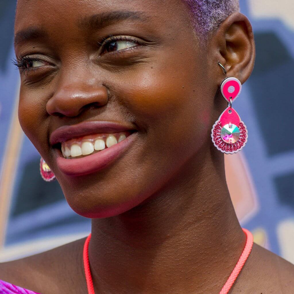 A close up of a smiling black woman's face. She is wearing a pair of small, neon pink dangly earrings and an orange cord necklace.