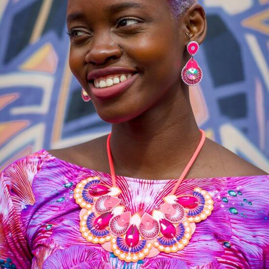 A close up of a young black woman wearing a bright pink and purple top, large orange bib necklace and pink earrings. She is smiling and looking away from the camera