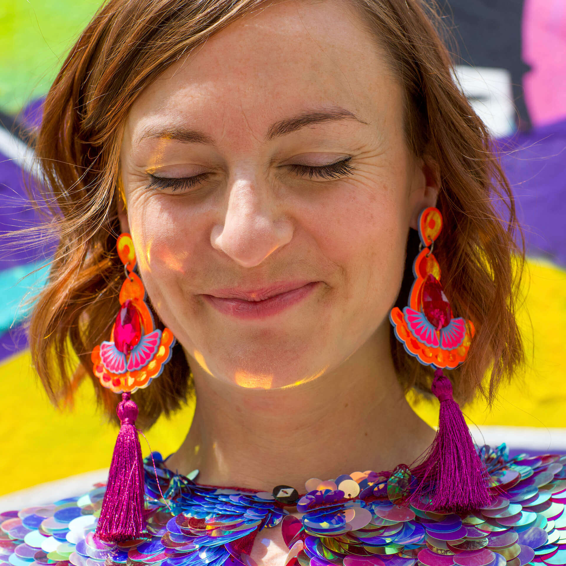 a white woman with short brown hair wearing large colourful tassel earrings is facing the camera with her eyes closed. The wall behind her is painted with bright colours and the image looks sunny and warm.