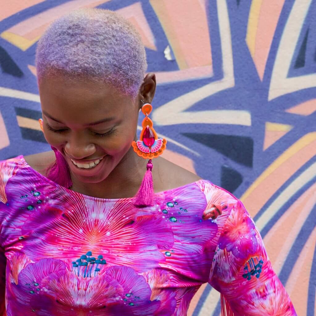 A young black woman with short lilac hair wearing a fitted, brightly coloured pink top and statement tassel earrings is looking down away from the camera and smiling. The wall behind her is painted with lilac and peach angular graffiti.