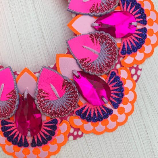 A close up of an ornately layered and printed Statement bib necklace. The orange, lilac pink and blue fabric layers are decorated with hot pink teardrop shaped jewels