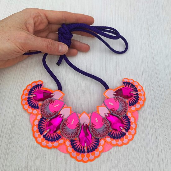 A ornate pink and orange statement jewel bib necklace is held by its dark purple cord ties by a woman's hand against an off white background
