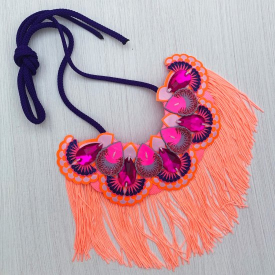 A statement jewel necklace with an ornately decorated bib in orange, pink, lavender and blue with a neon peach fringe and dark purple cord ties.