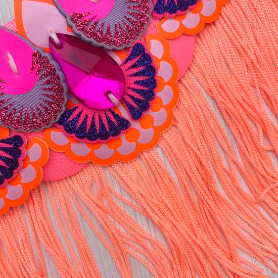A close up of a pink and orange bib necklace showing a hot pink cut glass jewel, glitter print fabric laser cut components and some neon pink fringe