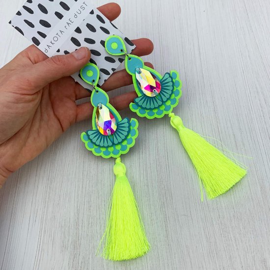 A pair of fluorescent yellow, lime and light blue jewel tassel earrings with iridescent teardrop shaped gems mounted on a black and white patterned, dakota rae dust branded card and held in a woman's hand against an off white background
