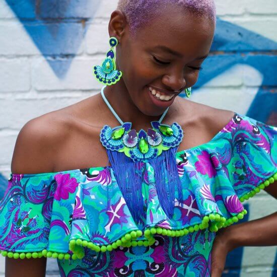 A young black woman with cropped lilac hair wears a pair of colourful, oversize statement earrings and a boldly patterned off the shoulder top. She is standing in front of a bright blue and white colourfully painted brick wall