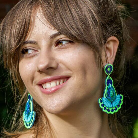 A young woman with brown fringe is smiling and gazing out to the left of the picture. She is wearing lime green and blue statement earrings with jewels