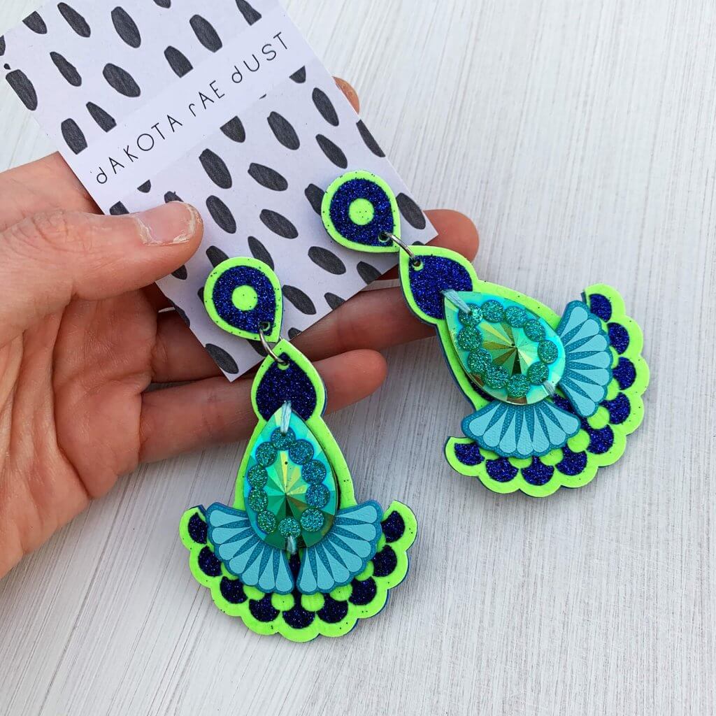 A pair of neon green and glittery blue teardrop and fan shaped jewelled earrings adorned with an glittery green gem are seen mounted on a black and white patterned, dakota rae dust branded card, held by a white hand against an off white background