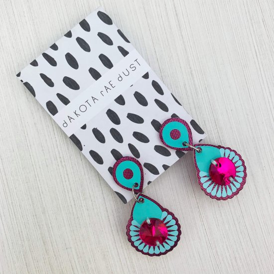 A pair of dangly mini jewel earrings in purple, turquoise and pink mounted on a black and white patterned, dakota rae dust branded card lie on an off white background