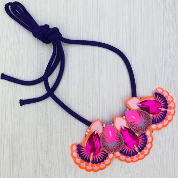 A colourful mini bib necklace in orange, pink, lilac and blue with purple cords lies on an off white background