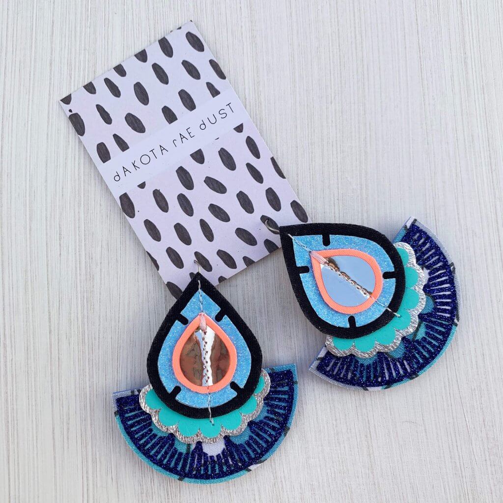 A pair a bright blue and turquoise oversize fan earrings on a black and white patterned dakota rae dust branded card, seen against a textured off white background