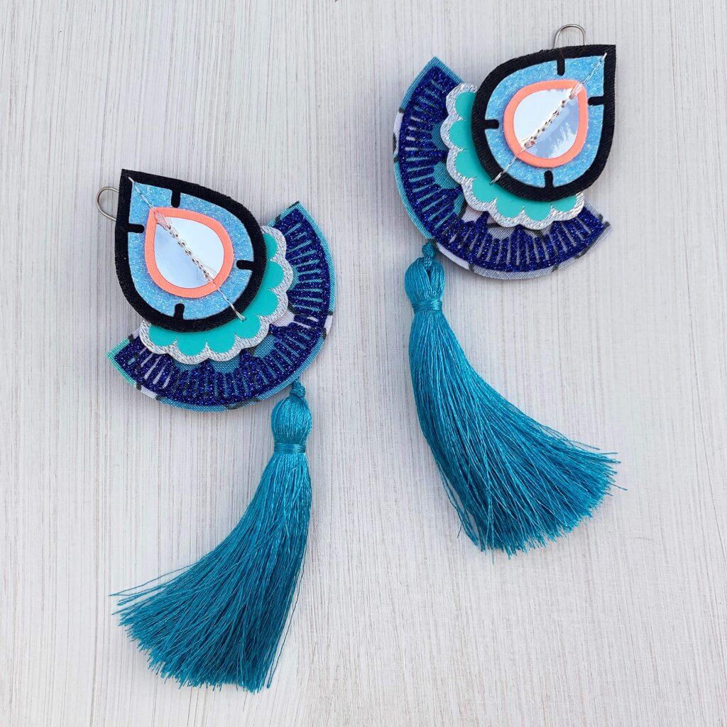 A pair of turquoise, blue, silver and coral large tasselled fan earrings lying side by side on an off white background. The silky turquoise tassels are curving in opposite directions.