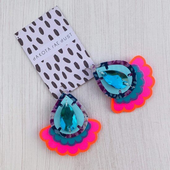 A colourful pair of teardrop shapes fluorescent pink, turquoise and blue oversize earrings featuring a bright scallop edged frill are seen displayed on a black and white dakota rae dust branded card, against a textured white background.
