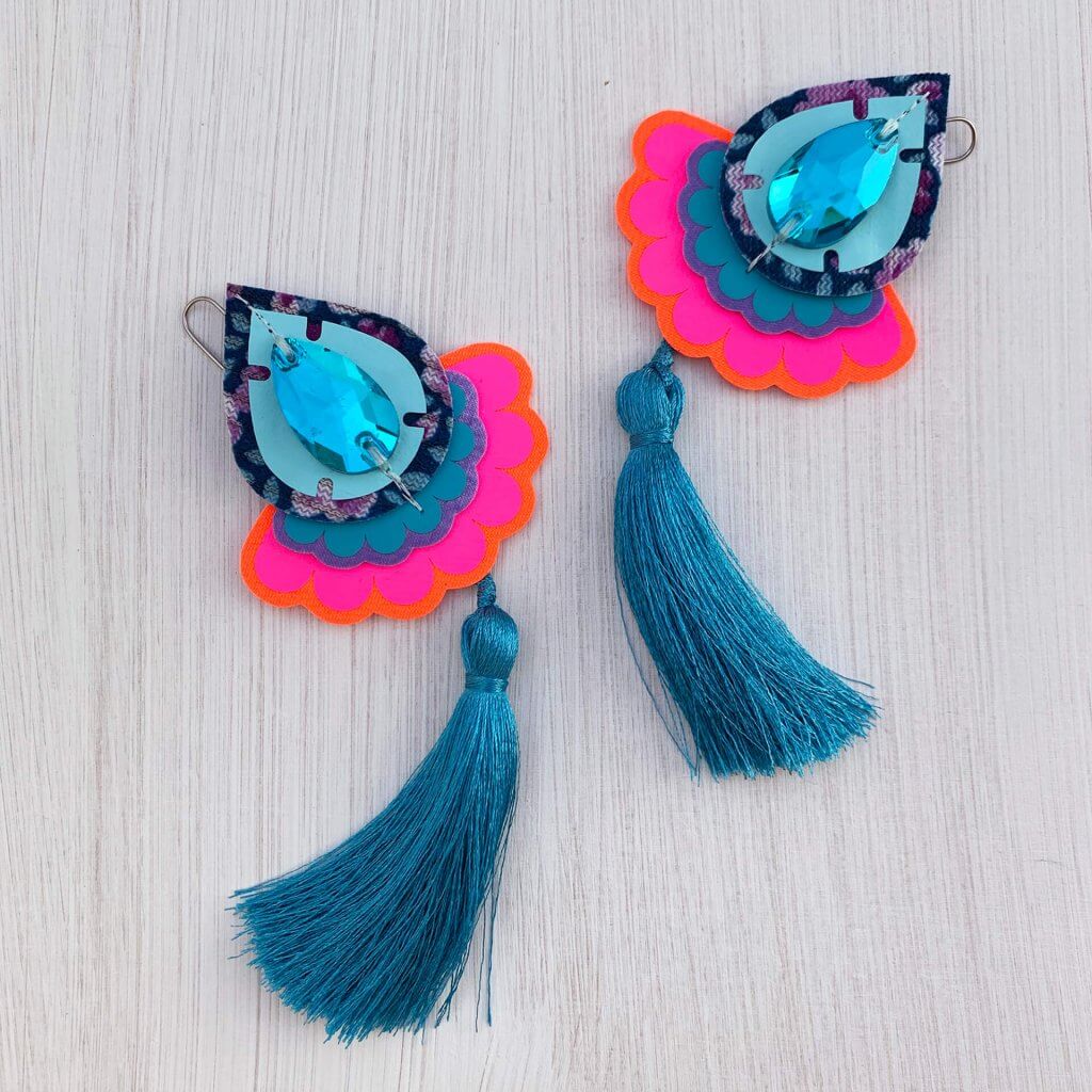 A pair of colourful teardrop shaped statement tassel earrings featuring turquoise jewels are seen lying on a textured white background. The earrings have turquoise tassels and fluorescent pink scallop edged trims.