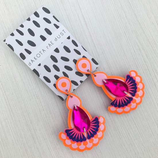 A pair of laser cut fabric Statement jewel earrings in orange, pink and blue mounted on a black and white patterned, branded dakota rae dust card lie on an off white background