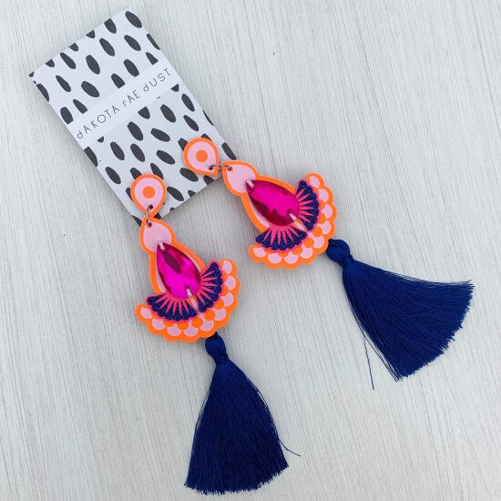 A pair of colourful Statement tassel earrings in orange with bright pink teardrop shaped jewels and deep blue tassels lying against on off white background