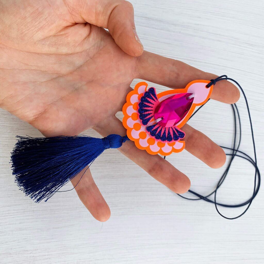 A orange, pink and blue tassel pendant necklace featuring a hot pink teardrop shaped jewel and thin blue cord is held in a woman's hand against an off white background