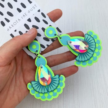 A pair of fluorescent yellow, lime and light blue teardrop jewel earrings with iridescent gems mounted on a black and white patterned, dakota rae dust branded card, held in a woman's hand.