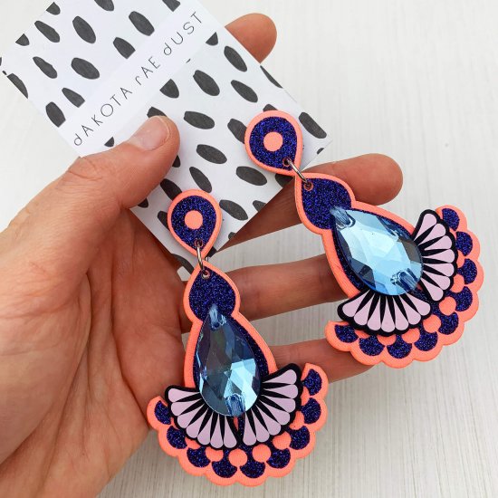 A pair of blue and peach teardrop jewel earrings mounted on a black and white patterned, dakota rae dust branded card are held in a woman's hand