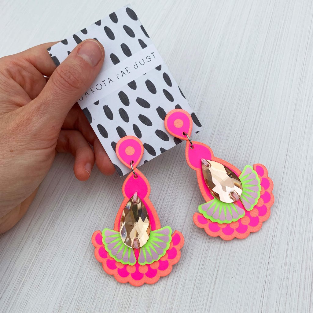 A pair of peach, pink and lime teardrop jewel earrings mounted on a black and white patterned, dakota rae dust branded card are held in a woman's hand against an off white background
