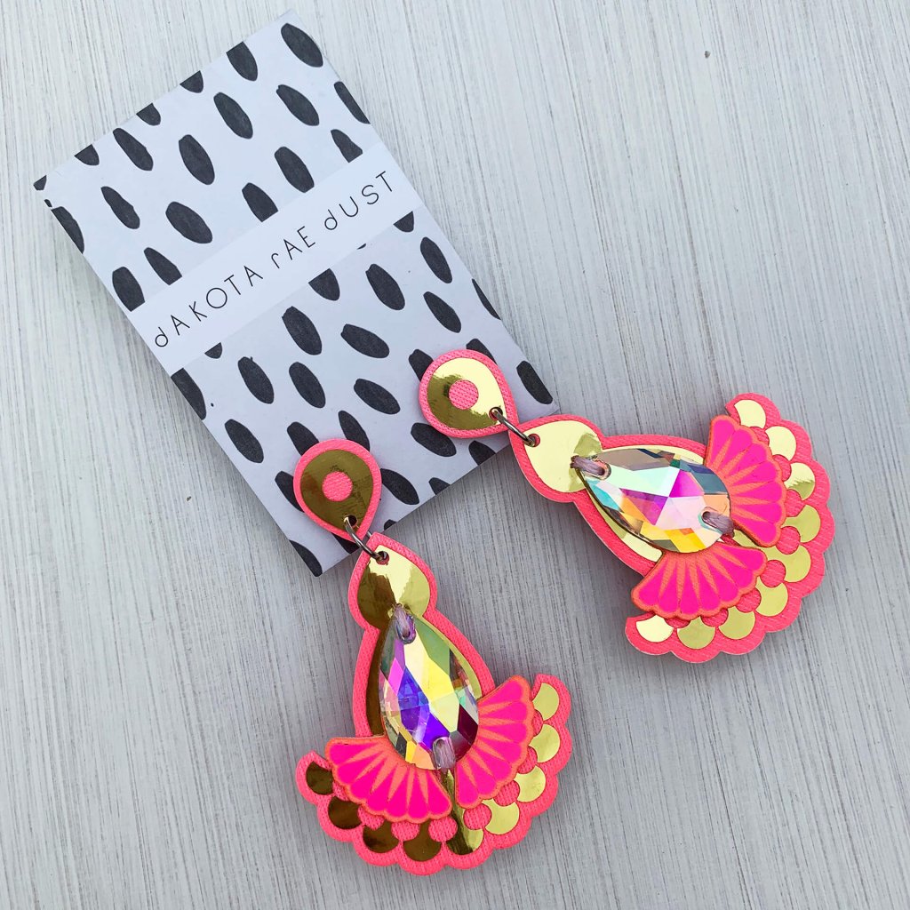 A pair of pink and gold mirrored teardrop jewel earrings adorned with iridescent gems mounted on a black and white patterned, dakota rae dust branded card are lying on an off white background