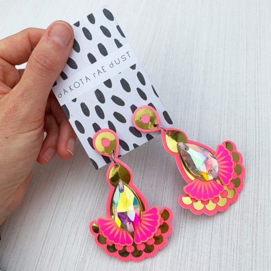 A pair of shiny gold and neon pink statement earrings with a teardrop shaped iridescent jewel, backed on a black and white patterned dakota rae dust branded card are held in a woman's hand against an off white background