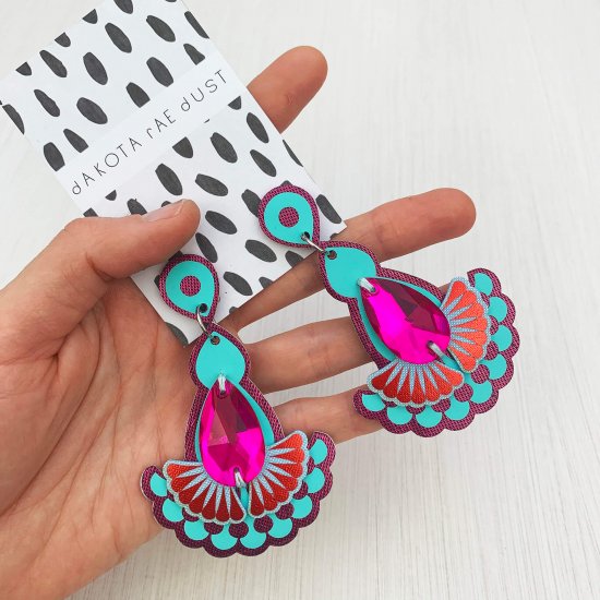 A pair of teardrop jewel earrings in purple, turquoise and pink mounted on a black and white patterned, dakota rae dust branded card and held in a woman's hand.