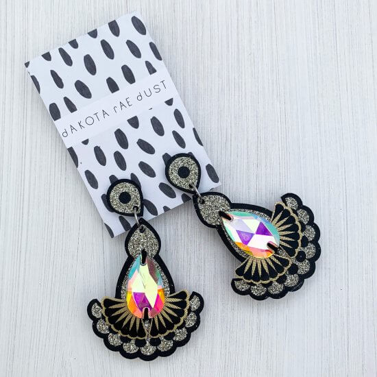 A pair of black, silver glitter and gold statement jewel earrings with iridescent teardrop shaped jewels, mounted on a black and white patterned, dakota rae dust branded card are seen against an off white textured background