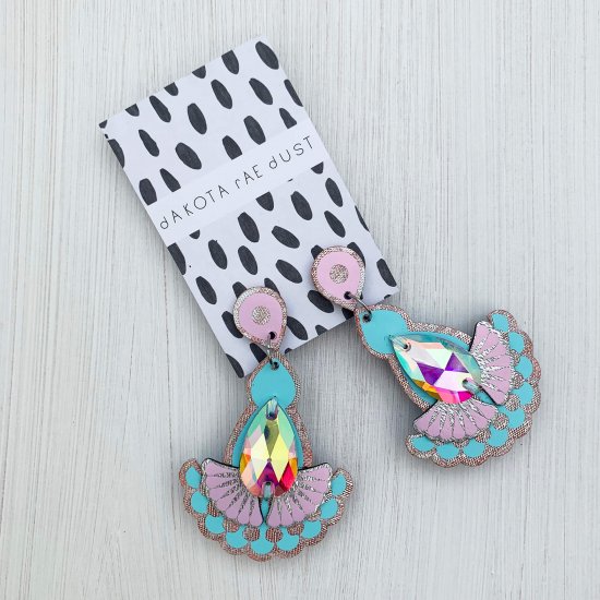 A pair of laser cut pastel pink and blue metallic statement earrings with teardrop shaped iridescent jewels, mounted on a black and white patterned, dakota rae dust branded card