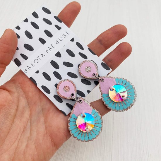 A pair of pastel pink and blue metallic mini jewel earrings mounted on a black and white patterned, dakota rae dust branded card are held in the open palm of a woman's hand